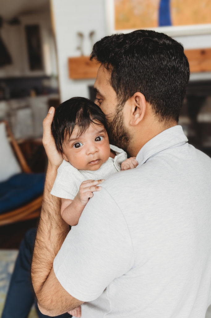 dad holding a newborn baby with open eyes looking into camera Indian newborn home session